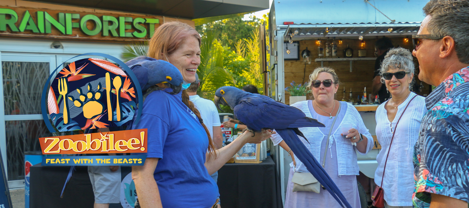 Zookeeper holding two Hyacinth Macaws while interacting with three guests at Zoobilee! Feast with the Beasts. Image contains Zoobilee event logo.