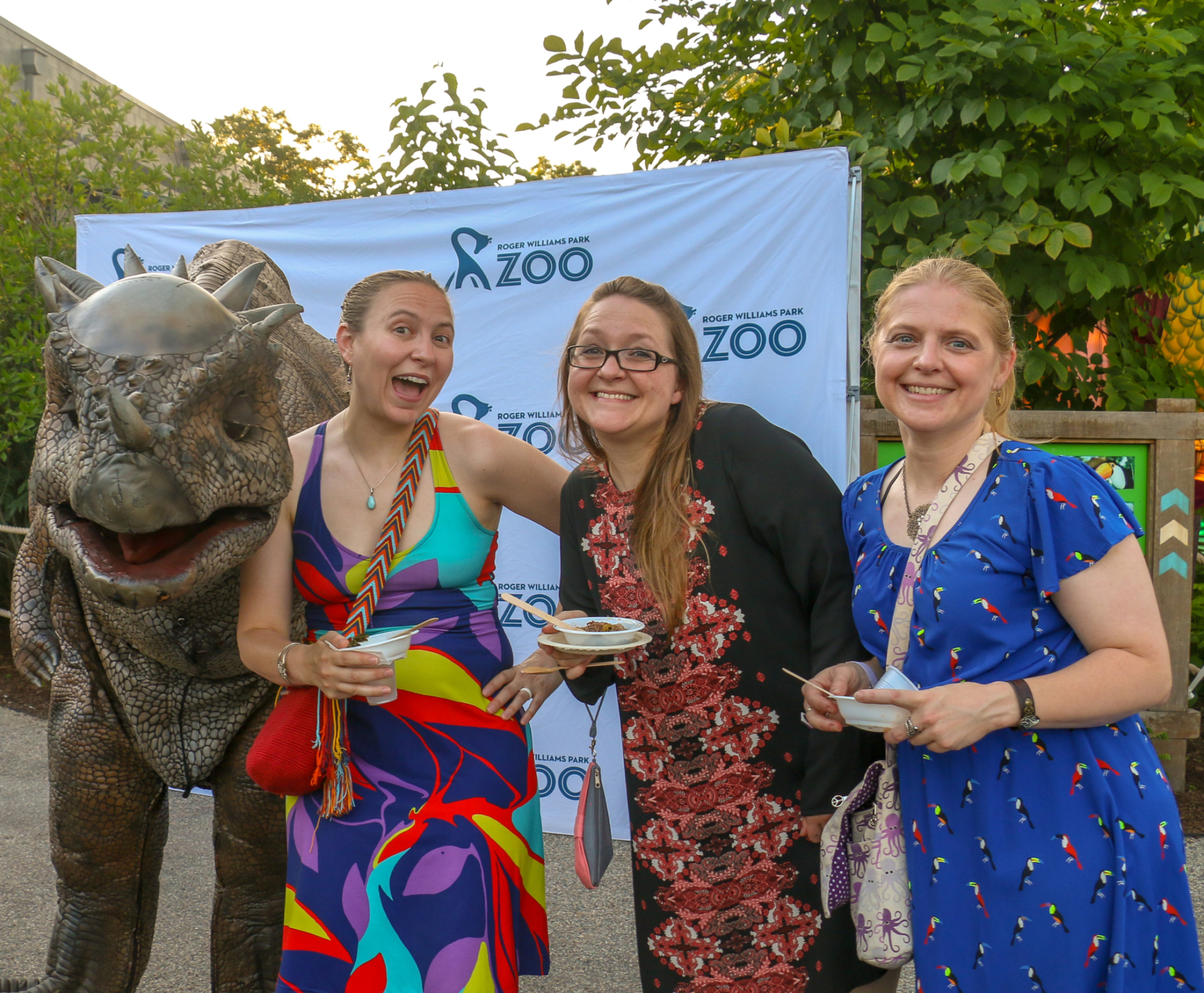 Three guests attending Zoobilee! Feast with the Beasts fundraiser posing with a dinosaur costume. Guests are smiling and holding samples of food from the restaurants participating in Zoobilee.