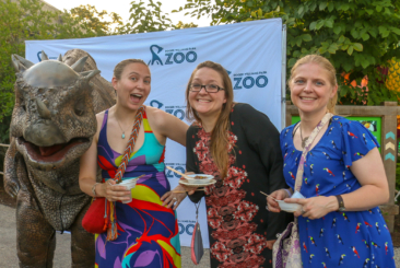 Three guests attending Zoobilee! Feast with the Beasts fundraiser posing with a dinosaur costume. Guests are smiling and holding samples of food from the restaurants participating in Zoobilee.