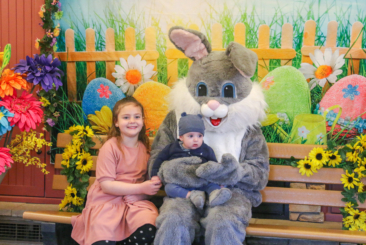 Two children posing and smiling with the Easter Bunny during an Easter Bunny meet and greet. Spring flowers and backdrop fill the background of the photo opportunity.