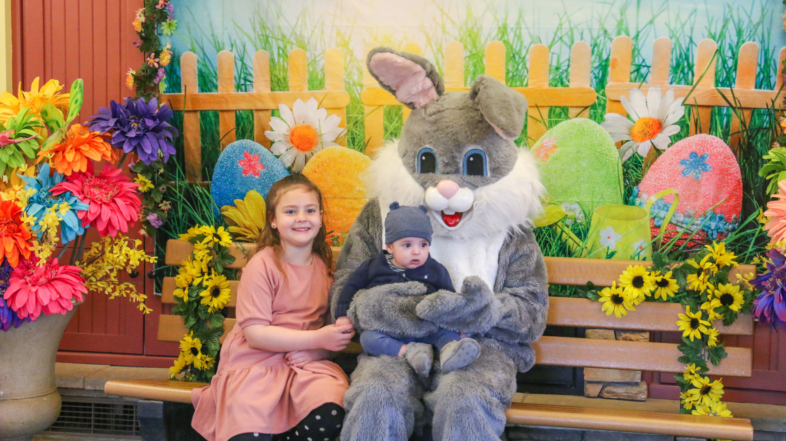 Two children posing and smiling with the Easter Bunny during an Easter Bunny meet and greet. Spring flowers and backdrop fill the background of the photo opportunity.