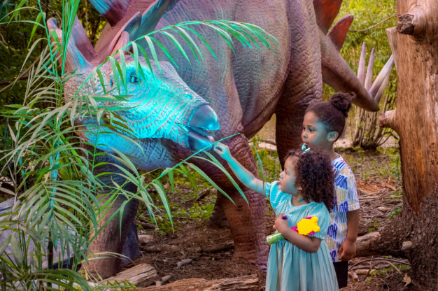 two children with an animatronic dinosaur lit up at dusk